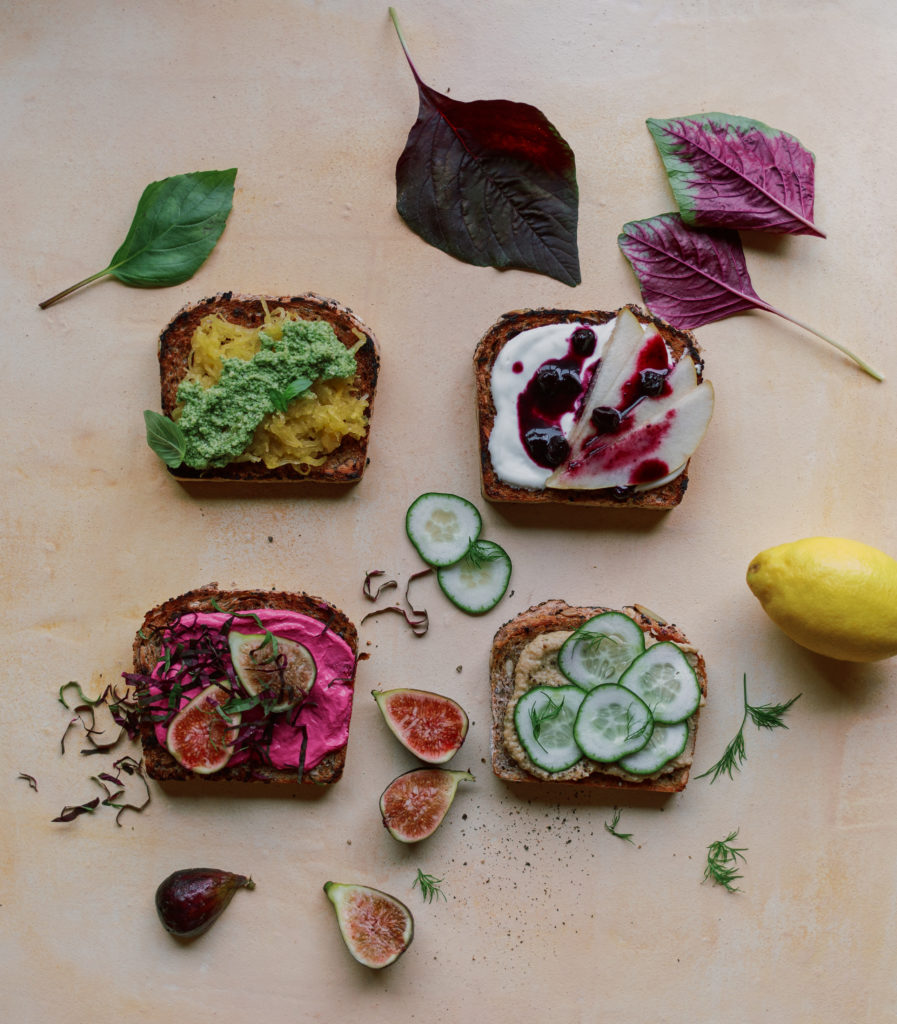 Open faced toasts on multi-seeded bread with various toppings, including hummus with lemon and dill, pink goat cheese, berry compote, and pesto.