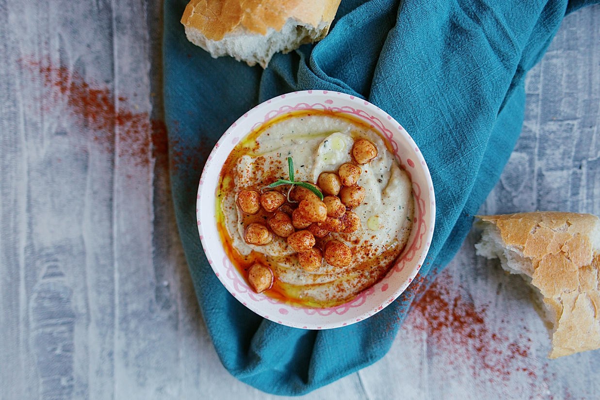Creamy and delicious dill hummus with a touch of lemon and paprika and garnished with olive oil and marinated chickpeas, resting on a blue towel and cement counter top.